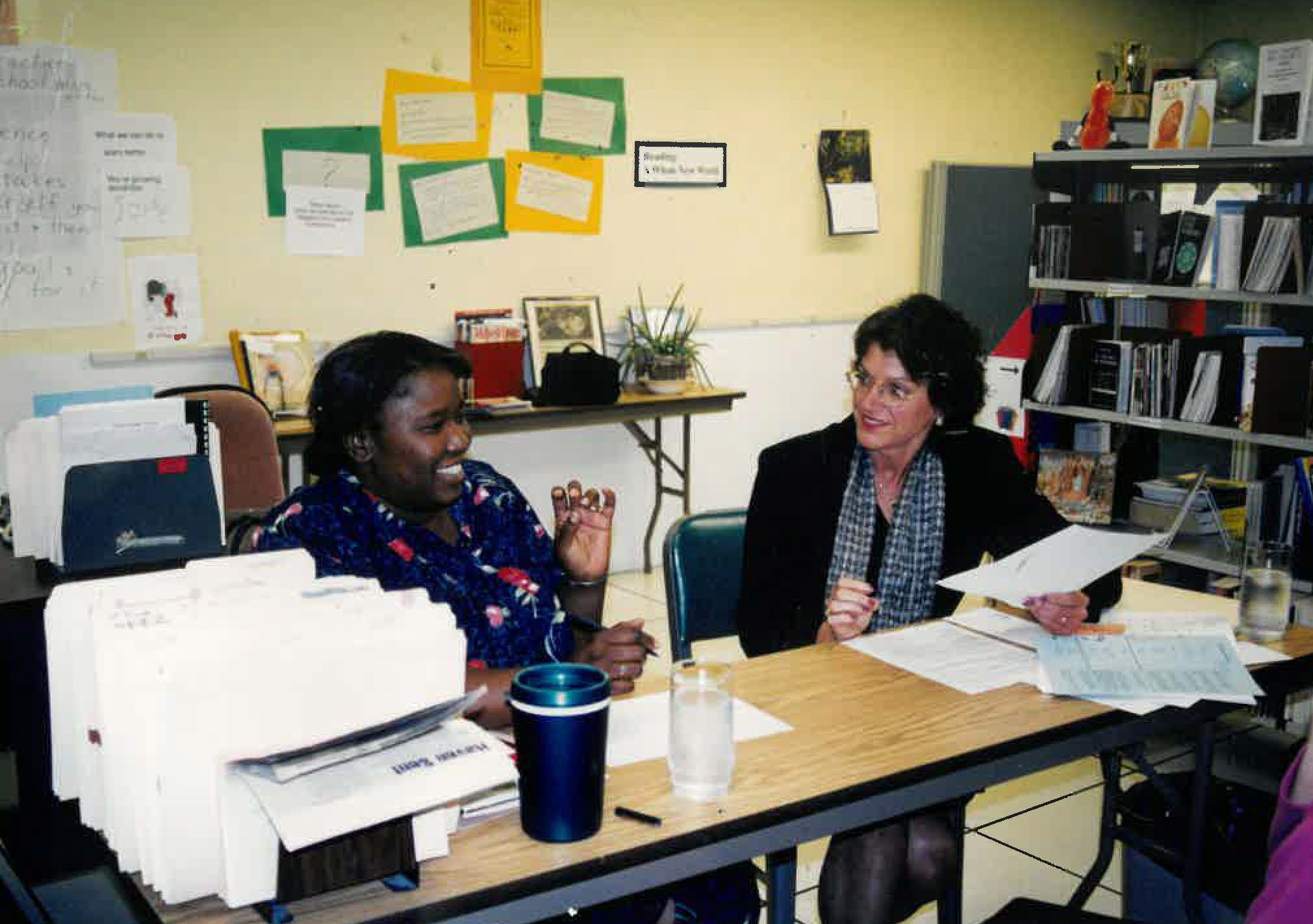 Two adults seated at a table in an office or classroom, reviewing papers together, with stacked documents, bookshelves, and posted notes visible in the room.