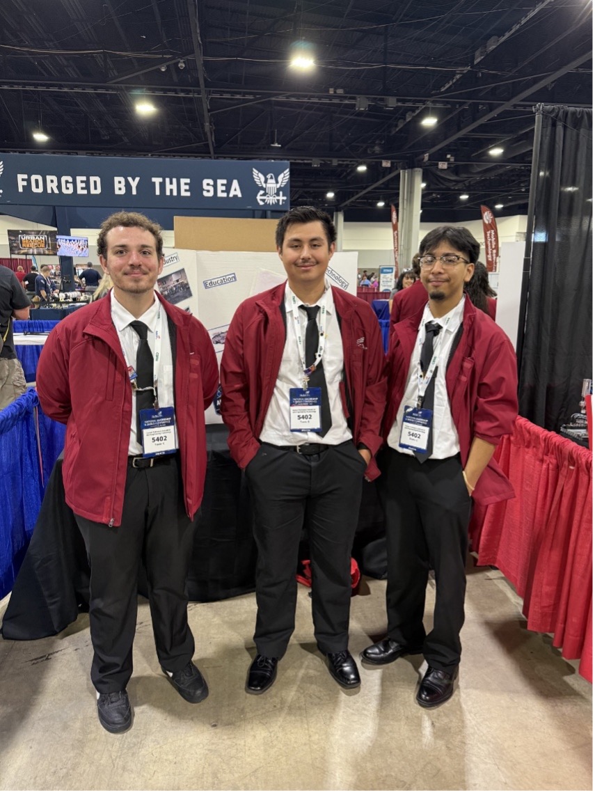 Three students wearing red suits stand in front of team display